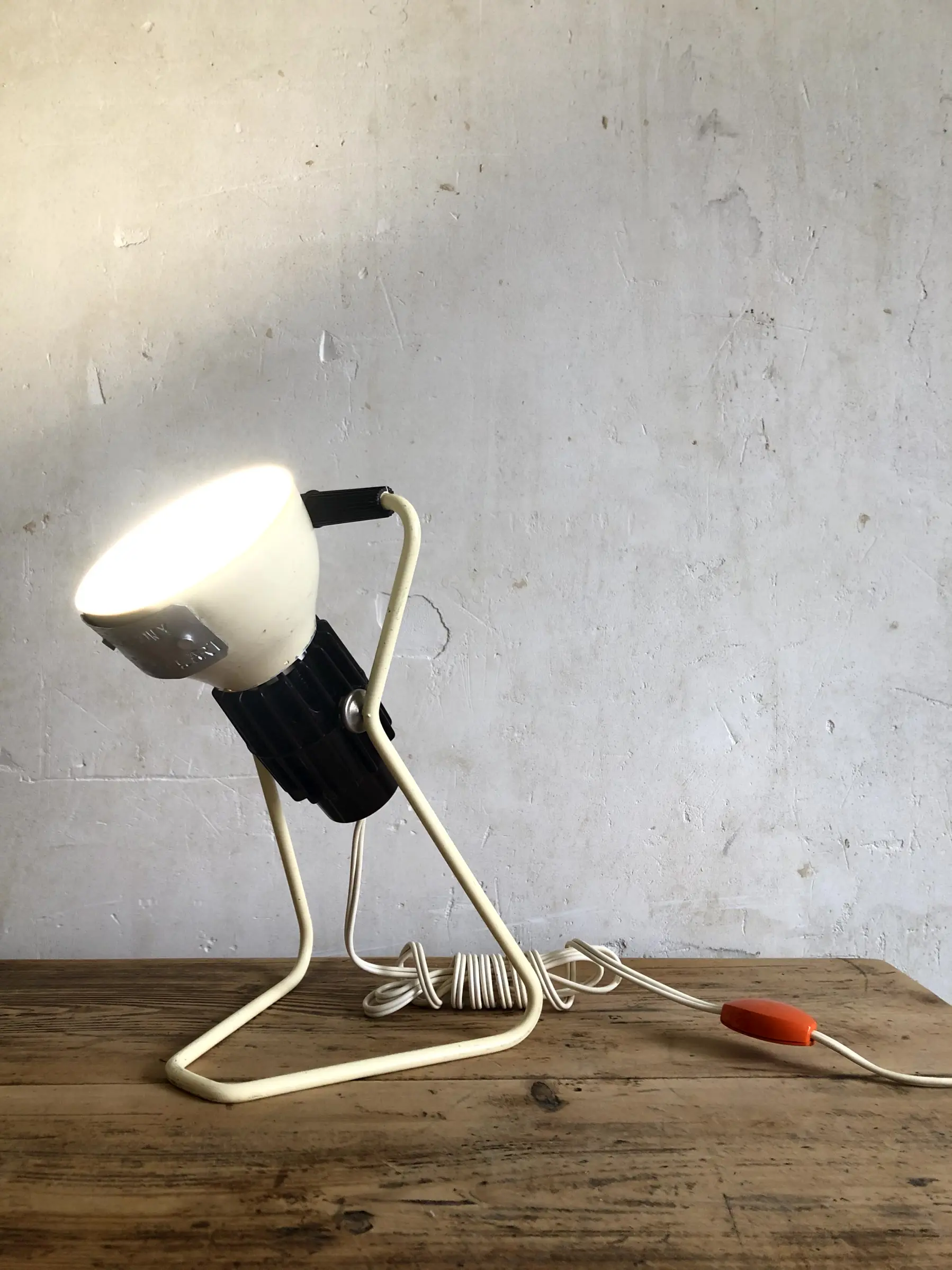 La lampe Omni allumée et posée sur un bureau, en position haute, avec son câble d'alimentation qui passe derrière le socle